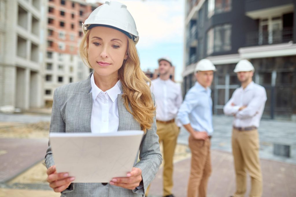 attentiveness-businesswoman-with-long-blond-hair-safety-helmet-looking-closely-document-standing-construction-site-working-group