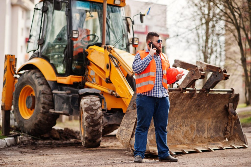 beard-worker-man-suit-construction-worker-safety-orange-helmet-sunglasses-against-tractor-with-mobile-phone-hand