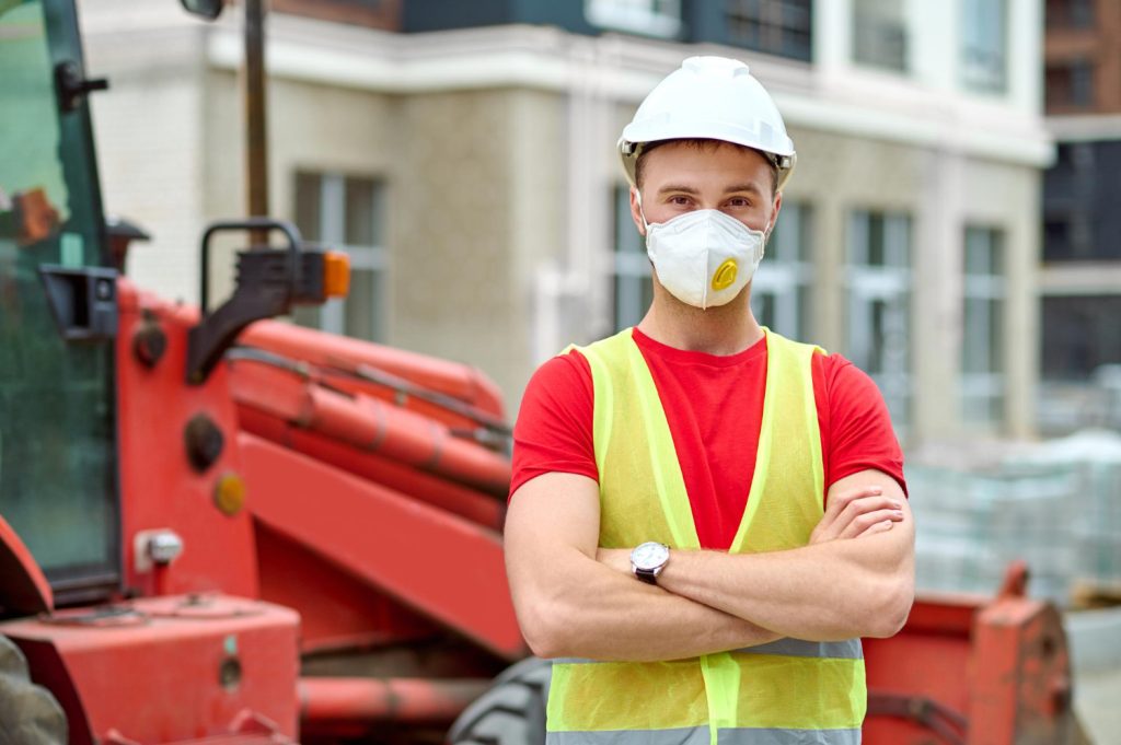 optimism-man-protective-mask-helmet-work-vest-with-folded-hands-looking-camera-good-mood-construction-site-afternoon