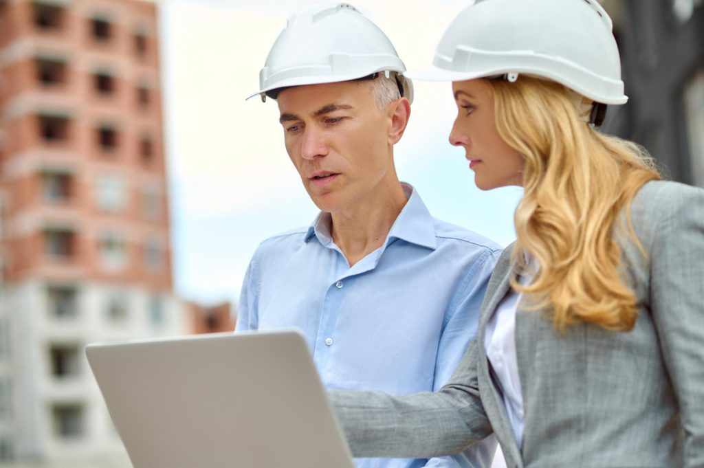 question-profile-demanding-business-woman-with-long-blond-hair-protective-helmet-looking-attentively-talking-man-with-laptop-standing-construction-site